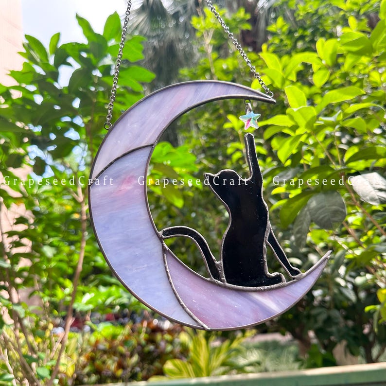 May include: Stained glass moon with a black cat silhouette reaching for a star. The moon is lavender and white, with a silver chain for hanging. The text "GrapeseedCraft" is visible.
