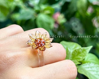 May include: A clear ring with a sunflower design. The ring has a transparent band and a flower with amber-coloured petals and a centre of small, reddish-brown beads. The ring is set against a blurred green background.