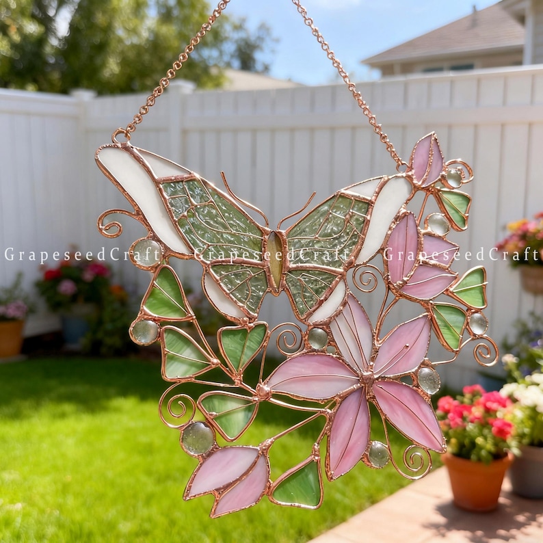May include: Stained glass butterfly and floral suncatcher with pink, green, and white glass. The butterfly has a green textured body and white wings. The flowers are pink and the leaves are green. The suncatcher is framed with copper wire.