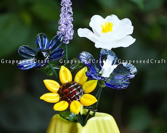 May include: A glass flower arrangement in a yellow vase. The arrangement includes a yellow sunflower with a brown center, blue and white flowers, and a purple lavender-like flower. The flowers are made of glass and have a translucent quality. The text "GrapeseedCraft" is visible.