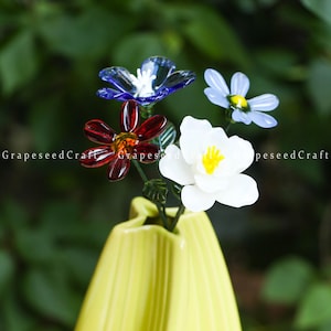 Puede incluir: Un jarrón amarillo contiene un ramo de flores de cristal en tonos rojos, azules y blancos. Las flores tienen pétalos detallados y tallos verdes. El fondo es un verde borroso.