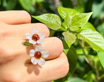 May include: A floral ring with two daisy-shaped flowers. The flowers have clear petals and amber centres. The ring has a gold-coloured band and a small green leaf accent. Green foliage forms the background.
