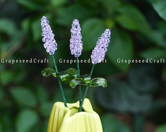 May include: Three lavender-colored, beaded flower stems with green leaves and stems are displayed in a yellow vase. The text "GrapeseedCraft" is visible on the image.