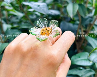 May include: A clear glass flower ring with a silver band. The flower has translucent petals with green edges and a yellow centre. The ring is held up by a hand against a blurred green background.