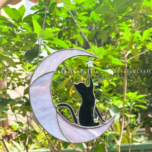 May include: A stained glass crescent moon with a black cat silhouette reaching for a star. The moon is a mix of white and lavender glass, and the star is clear. The piece hangs from a silver chain.