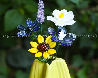May include: A vibrant glass flower arrangement in a yellow vase. The bouquet includes a yellow sunflower, blue and white flowers, and a purple lavender-like stem. The text "GrapeseedCraft" is visible on the image.