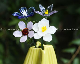 May include: A yellow vase holds four glass flowers in shades of blue, purple, white, and pink. The flowers have detailed petals and yellow centers. The vase is a light yellow color and has a ribbed texture.