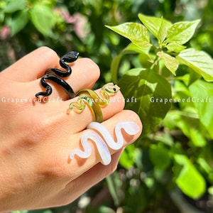 Op de afbeelding: Drie glazen slangenringen in zwart, groen en wit worden op een hand getoond. De zwarte ring is glanzend, de groene ring heeft een gele tint en de witte ring is doorschijnend. De ringen bevinden zich in een natuurlijke omgeving met groen gebladerte.