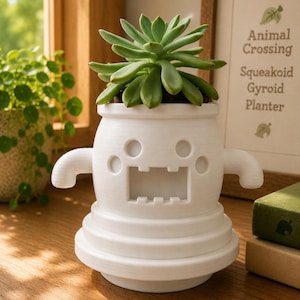 Peut inclure: Un pot de fleurs blanc imprimé en 3D en forme de personnage Squeakoid Gyroid d'Animal Crossing. Le pot présente une base large et étagée, deux petits bras latéraux, une bouche rectangulaire et des yeux circulaires. Une plante succulente verte est plantée à l'intérieur.