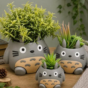 May include: Three grey ceramic planters shaped like cartoon creatures with round white eyes, black whiskers, and tan bellies with dark markings. Each planter holds a different green plant, ranging from a bushy shrub to spiky succulents.