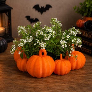 May include: A cluster of four bright orange, ribbed pumpkin-shaped planters arranged in a circle. Each pumpkin features a distinct vertical texture and a central opening at the top, currently holding a lush arrangement of small white flowers.