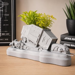 Könnte beinhalten: Ein hellgrauer, 3D-gedruckter Übertopf in Form eines AT-AT-Läufers aus Star Wars, mit rechteckigem Körper, mechanischen Beindetails und flacher Basis. Der Übertopf enthält eine leuchtend grüne Kunstpflanze mit kleinen, spitzen Blättern.