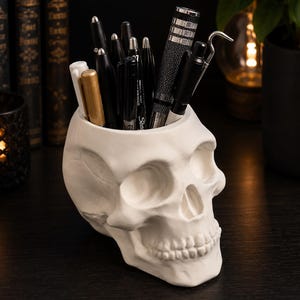 May include: A matte white skull-shaped desk organizer holding a collection of various pens and markers. The hollow cranium serves as a container for writing instruments, including black, gold, and white pens, along with a metallic textured pen.