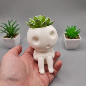 Könnte beinhalten: Weißer Keramik-Pflanztopf in Form einer Person mit einer kleinen Sukkulentenpflanze im Kopf. Der Topf hat zwei kleine Augen und einen Mund.