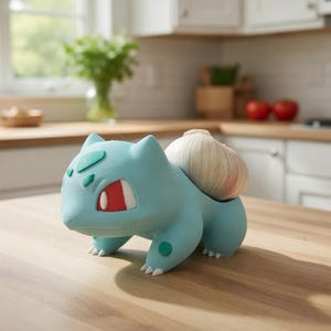 May include: A light blue Bulbasaur-shaped garlic keeper with a garlic bulb inside. The kitchen decor item is on a wooden countertop. The Bulbasaur has green spots and red eyes. The background includes a window, tomatoes, and a stove.