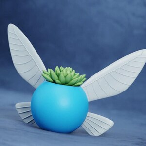 May include: A bright blue spherical planter featuring four white, wing-shaped attachments extending from the sides. A small, light green succulent plant with pointed, fleshy leaves is potted inside the top opening of the blue container.