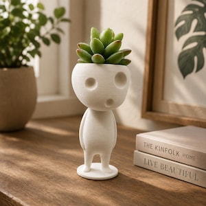 May include: A white, matte-finished planter shaped like a stylised humanoid figure with a large, rounded head and two circular eye-like indentations. A small, green succulent plant is potted inside the head of the figure, which stands on a circular base.