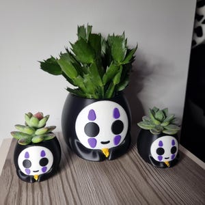 Puede incluir: Tres macetas de cerámica negra y blanca con forma del personaje Sin Cara de la película de animación El viaje de Chihiro. Las macetas están llenas de pequeñas suculentas verdes.