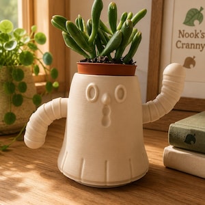 Peut inclure: Un cache-pot en céramique couleur crème en forme de fantôme avec deux bras levés, une expression faciale surprise et des lignes verticales à la base. Le pot contient une plante succulente verte dans un petit pot en plastique marron.