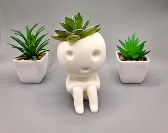May include: White ceramic planter shaped like a person with a small succulent plant in the head. The planter is sitting on a gray surface.