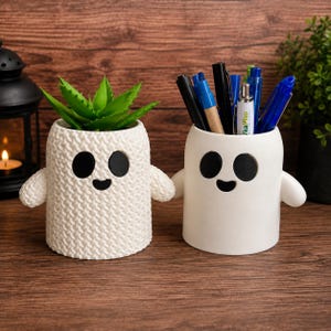 May include: Two white ghost-shaped containers with black oval eyes and mouths. The left container has a textured knit-like surface and holds a green succulent plant. The right container has a smooth surface and holds various pens and markers.