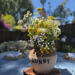 May include: A round, two-toned tan and brown ceramic pot with the word HUNNY printed in black letters. The pot features a yellow drip design at the rim and is filled with a bouquet of yellow and white flowers, including a small tiger character topper.