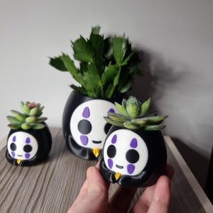 May include: Three black and white planters shaped like a character with a white face, black eyes, and purple markings. Each planter holds a green succulent plant. The planters have a smooth finish and a small yellow detail at the base.