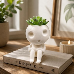 May include: A white, matte-finished planter shaped like a stylised, seated humanoid figure with a round head, hollow eye sockets, and a small mouth. The planter holds a vibrant green succulent plant with thick, layered leaves, resting on a book.