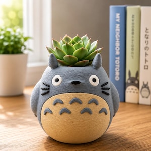 May include: A gray and beige character-shaped planter featuring a rounded body, two pointed ears, white eyes with black pupils, and black whiskers. A green succulent plant is potted inside the top. The planter has a textured surface.