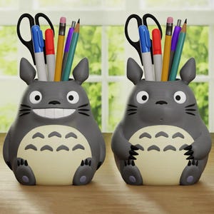 Puede incluir: Dos organizadores de escritorio grises con forma del personaje Totoro, cada uno con bolígrafos, lápices y tijeras. Los organizadores tienen una barriga beige claro y una cara sonriente. El fondo muestra una ventana con follaje verde.
