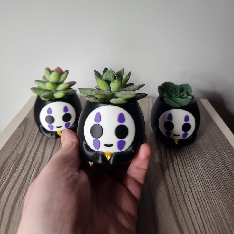 May include: Three black, rounded planters featuring a white mask design with purple accents and a small yellow detail at the base. Each planter holds a green succulent plant. The design mimics a character face with black eyes and a small mouth.