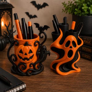 May include: Two cylindrical pen holders featuring Halloween designs. One is orange with a black jack-o'-lantern and ghost motif, while the other is a black and orange swirling ghost shape. Both holders contain various black and orange pens.