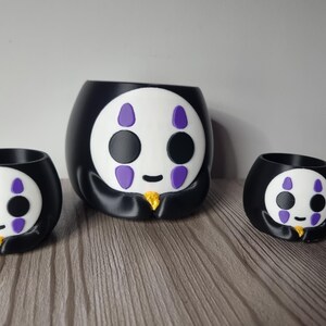 May include: A set of three black and white planters featuring a stylized character face with purple markings above and below the eyes, a small black mouth, and a gold-colored accent at the base, arranged in a group of one large and two smaller sizes.