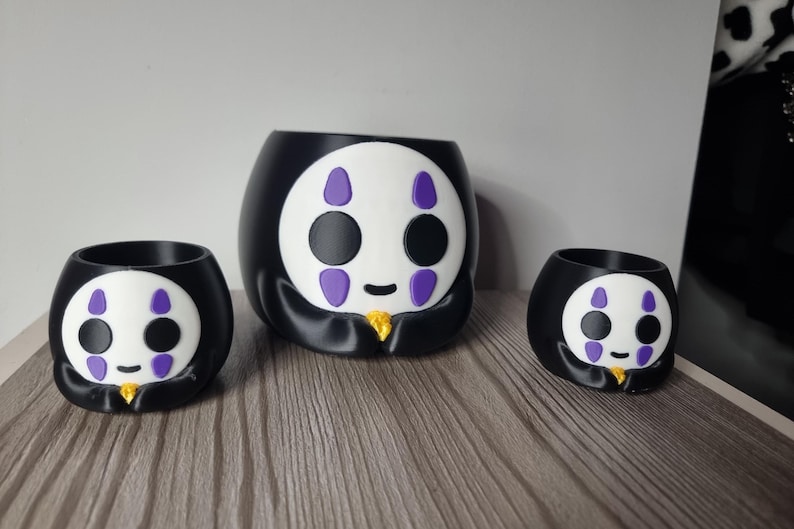 May include: A set of three black and white planters featuring a stylized character face with purple markings above and below the eyes, a small black mouth, and a gold-colored accent at the base, arranged in a group of one large and two smaller sizes.