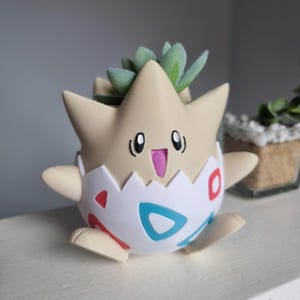 May include: A Togepi-shaped planter, featuring a light tan head and arms, and a white and patterned eggshell body with red and blue accents. The planter holds a small succulent plant. The design is inspired by the popular anime series.