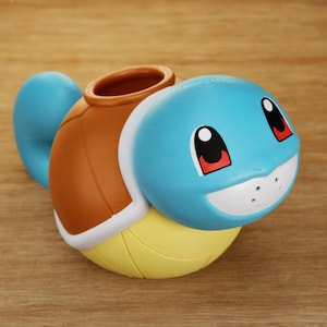 May include: A 3D-printed Squirtle-shaped container. The container features a light blue head with black and red eyes, a white mouth, and a brown shell. The base is yellow, and a blue tail extends from the back.