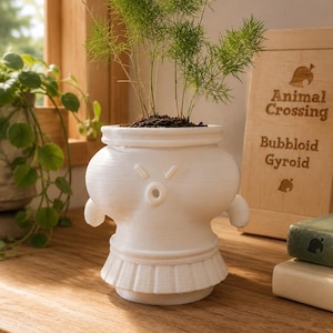 Peut inclure: Un pot de fleurs blanc imprimé en 3D en forme de Bubbloid Gyroïde d'Animal Crossing, avec un corps arrondi, de petits bras et une base plissée. Le pot contient une fougère asperge verte et est placé près d'un panneau en bois avec le texte Animal Crossing Bubbloid Gyroid.