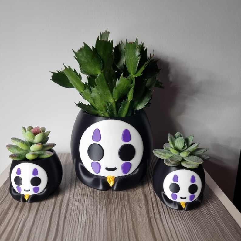 May include: Three black and white planters shaped like a character with a white face, purple markings, and a small gold accent. Each planter holds a green succulent plant, with the central planter being the largest of the three items.