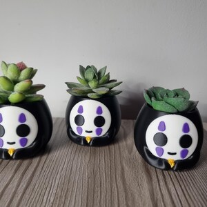 May include: Three black and white planters shaped like a character with a white face, purple markings, and a small yellow accent at the base. Each planter holds a green succulent plant with thick, fleshy leaves arranged in a rosette pattern.