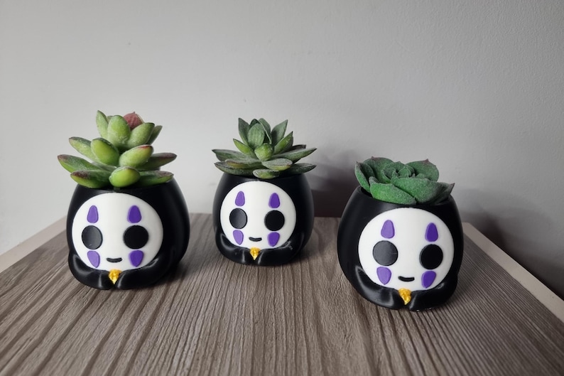 May include: Three black and white planters shaped like a character with a white face, purple markings, and a small yellow accent at the base. Each planter holds a green succulent plant with thick, fleshy leaves arranged in a rosette pattern.