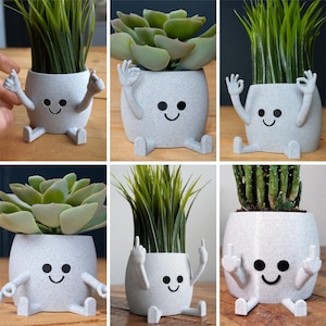 May include: A collection of gray, smiling character planters with various types of greenery. Each planter has arms and legs, and the faces have black eyes and a curved smile. The plants include succulents, grass, and a cactus.