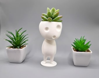 May include: White ceramic planter shaped like a person with a small succulent plant in the head. The planter is standing on a gray surface.