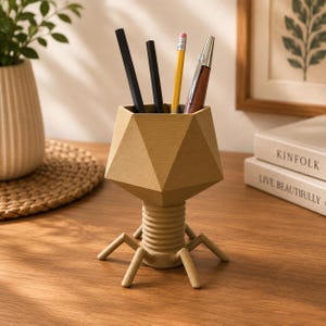 May include: A tan, geometric, bacteriophage-shaped pencil holder with a faceted head, a ribbed cylindrical neck, and six thin, angled legs. The holder contains two black pens, one yellow pencil, and one silver and wood-toned pen.