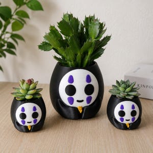May include: Three black and white planters shaped like a character with a white face, purple markings, and black eyes. The planters feature a small yellow detail at the base and hold green succulent plants of varying sizes in a rounded design.
