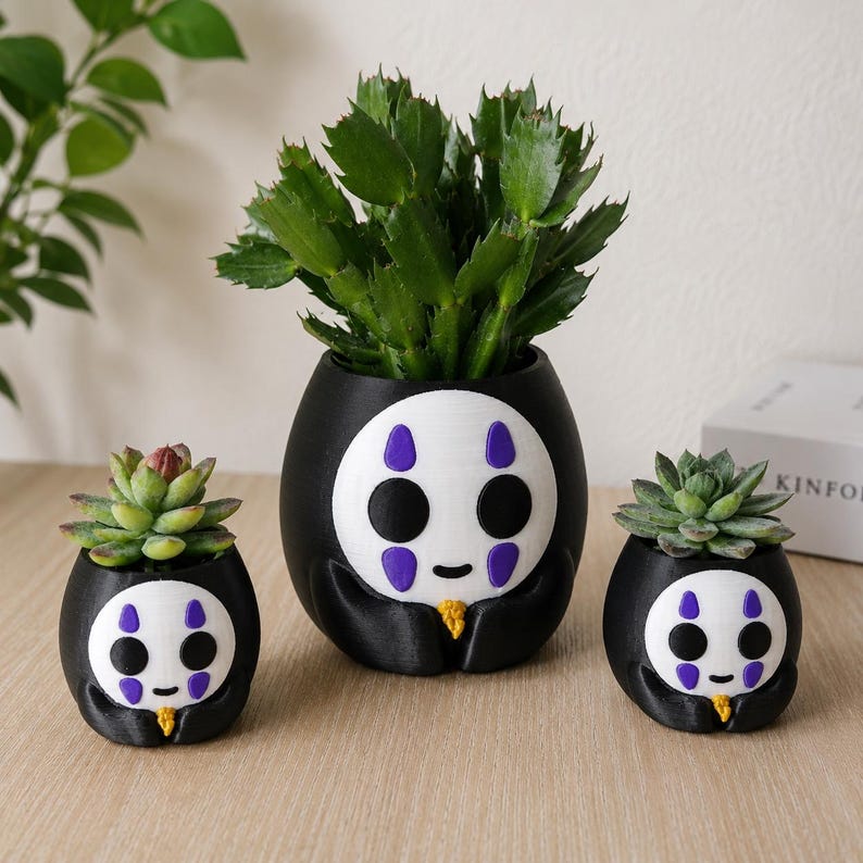 May include: Three black and white planters shaped like a character with a white face, purple markings, and black eyes. The planters feature a small yellow detail at the base and hold green succulent plants of varying sizes in a rounded design.