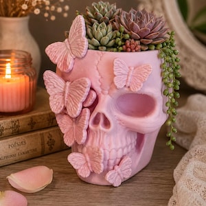 May include: A light pink skull-shaped planter adorned with several raised butterfly figures on its surface. The planter contains a variety of green and purple succulent plants, including a trailing string of pearls plant hanging over the side.
