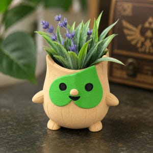 Peut inclure: Un pot de fleurs beige, texturé et arrondi, en forme de créature avec un grand masque en forme de feuille verte couvrant son visage. Le pot présente de petits bras et pieds, et il est rempli d'herbe verte artificielle et de brins de lavande.