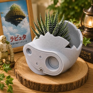 May include: A light grey, textured planter shaped like a robotic head with a jagged, broken-egg top edge. A green, white-spotted succulent plant grows from the centre. The planter features circular indentations resembling eyes and mechanical details.