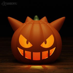 May include: A matte orange pumpkin-shaped lamp featuring a Gengar-inspired design with two pointed ears, a wide grinning mouth, and sharp triangular eyes. The interior glows with a warm yellow light, illuminating the cut-out facial features.