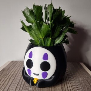 May include: A black and white planter shaped like a character with a white face, black circular eyes, and purple teardrop markings above and below the eyes. The character holds a small yellow object. A green succulent plant grows from the top.